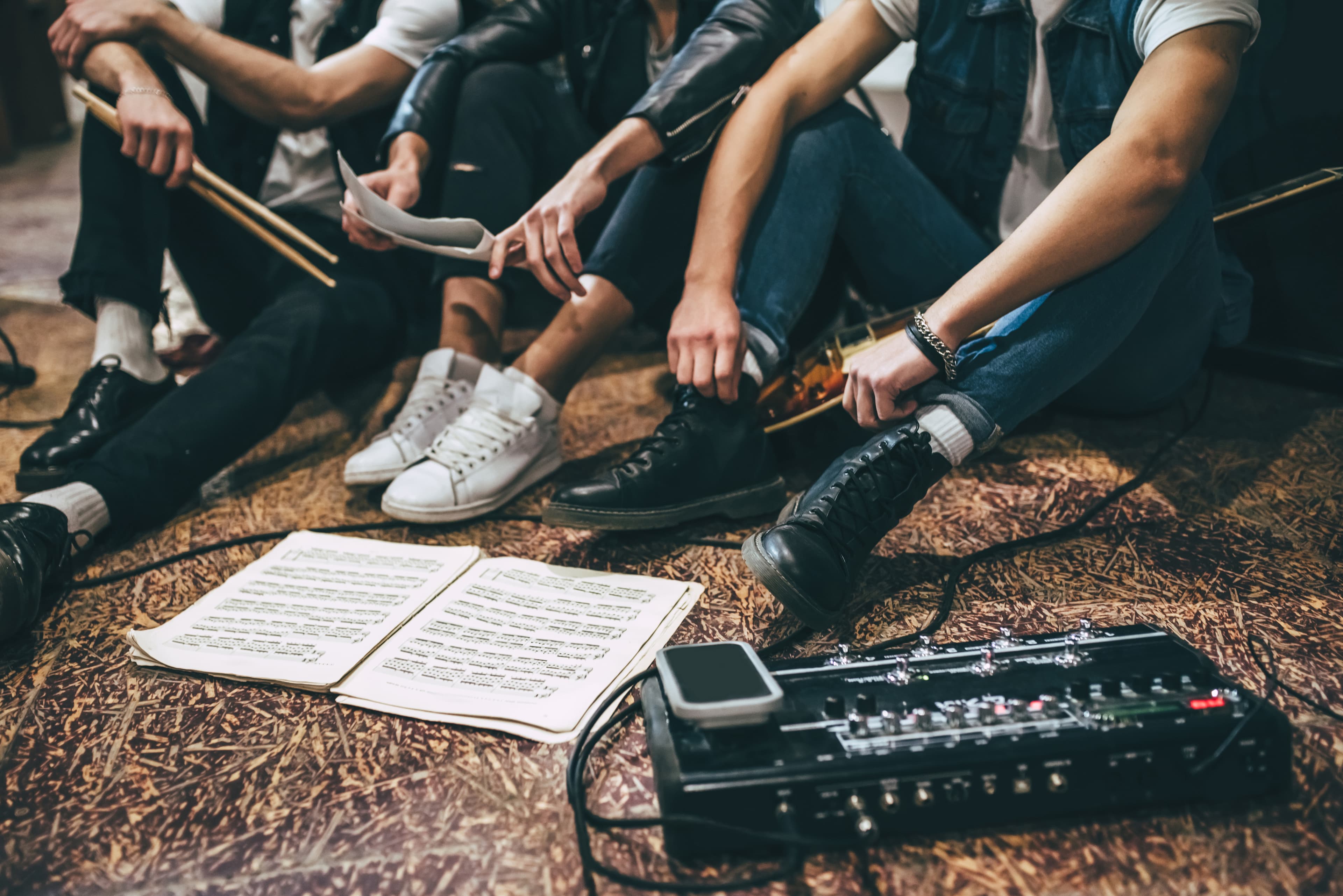 A group of musicians sitting on the floor with musical instruments, sheet music, and equipment during a practice session.