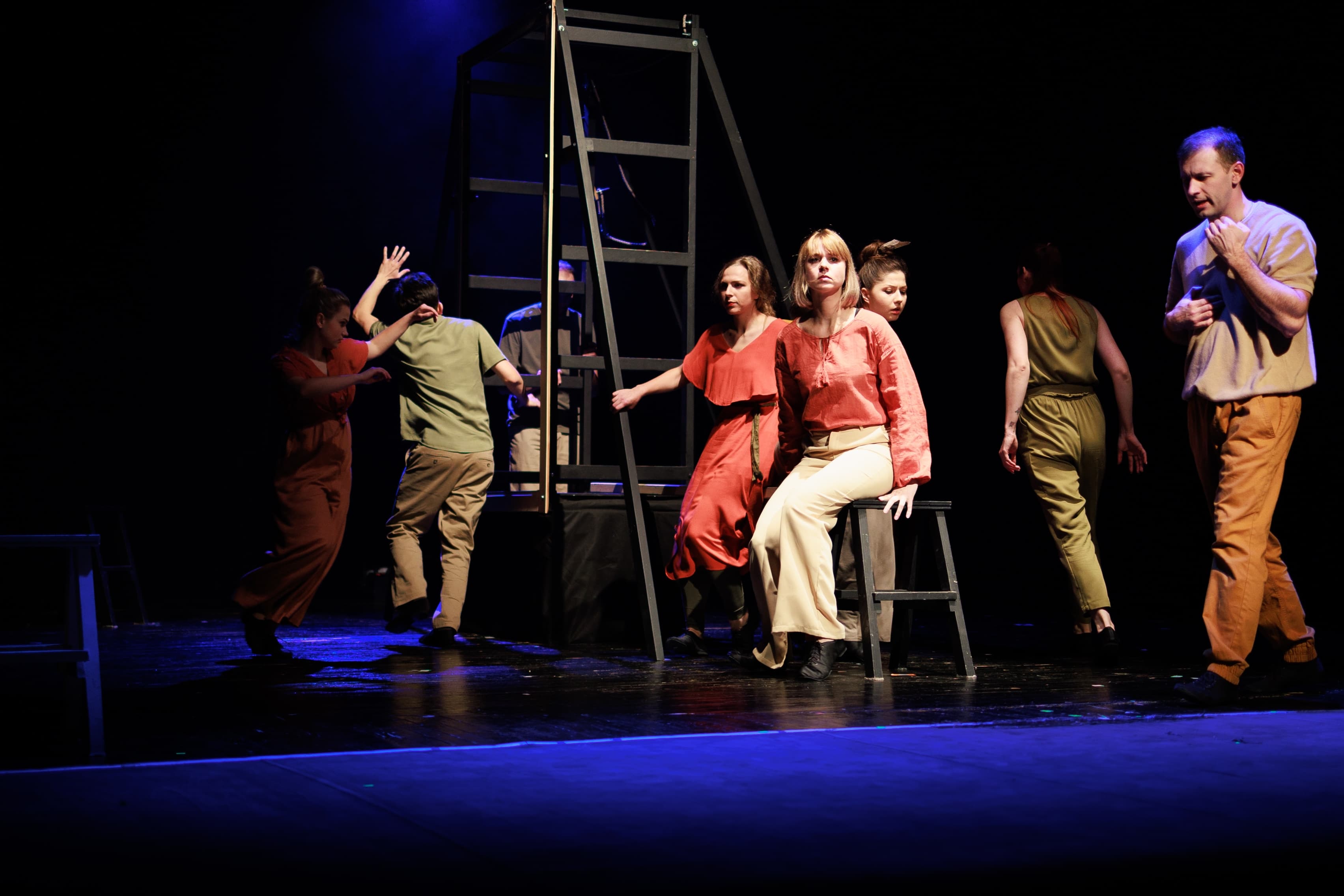 Several performers on stage under dramatic lighting, with a prominent black ladder structure in the background. The performers are dressed in simple, earthy-toned costumes, and their poses suggest movement and interaction, creating a dynamic theatrical scene.
